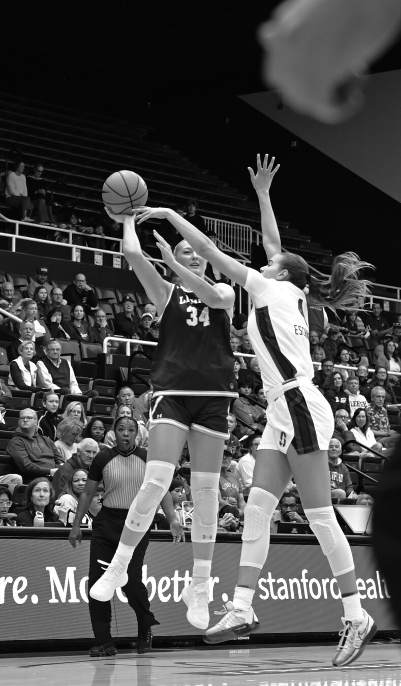 Action shot of two basketball players in the air. The one in black on the left has the ball and looks to be shooting or passing. The player in white on the right is trying to block the ball.