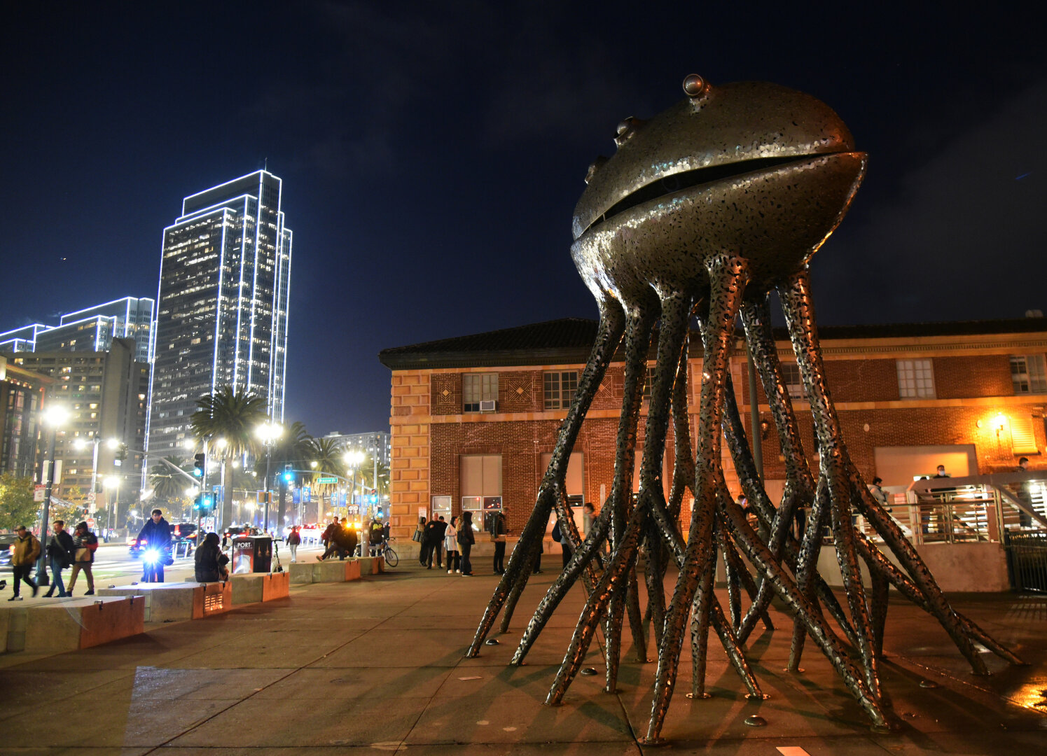 a tall statue of a many tentacled creature with a clam like mouth sits on the side of a well lit city street