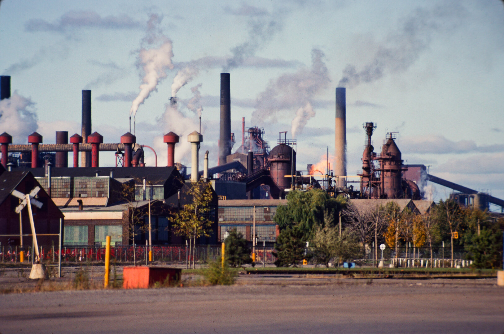 colour photo of active industrial building skyline including several smokestacks with plumes. parking lot and trees in foreground