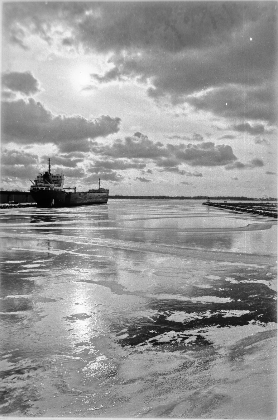 black and white photo of an ice covered ship channel, reflecting the clouds above. Cargo ship in background.
