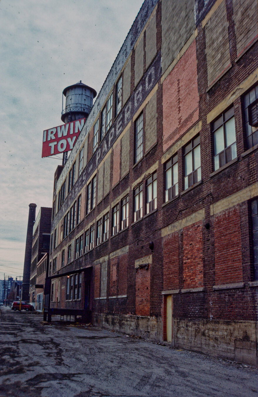 colour photo looking along side of large historic industrial building. smokestack in distance. water tower above along with sign reading "IRWIN TOY"