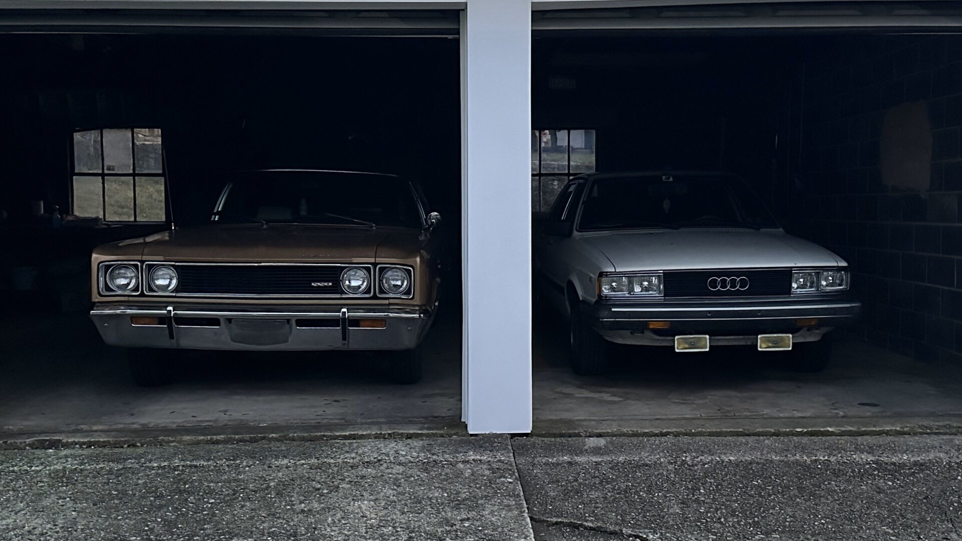 Photo of two cars in a garage. On the left is my 1969 AMC Rebel SST Sedan, which is two-tone brown. On the right is my 1982 Audi 4000 S Sedan, which is white, and has yellow Hella fog lights under the front bumper.