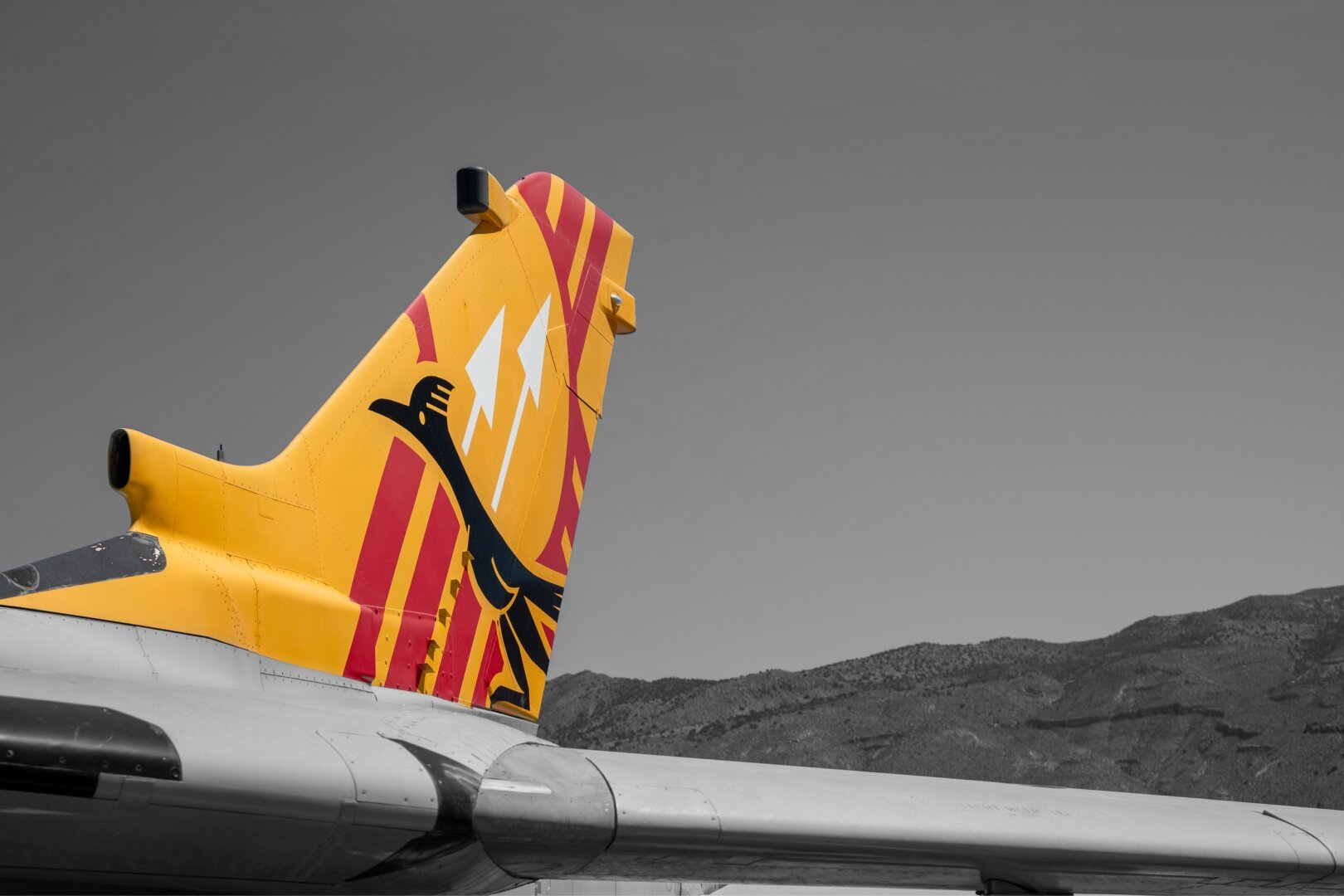 Close-up of a jet’s tail fin with a vivid red and yellow livery depicting a black Thunderbird and two white lightning bolts. The background is desaturated, showing a grayscale mountain range under a clear sky.