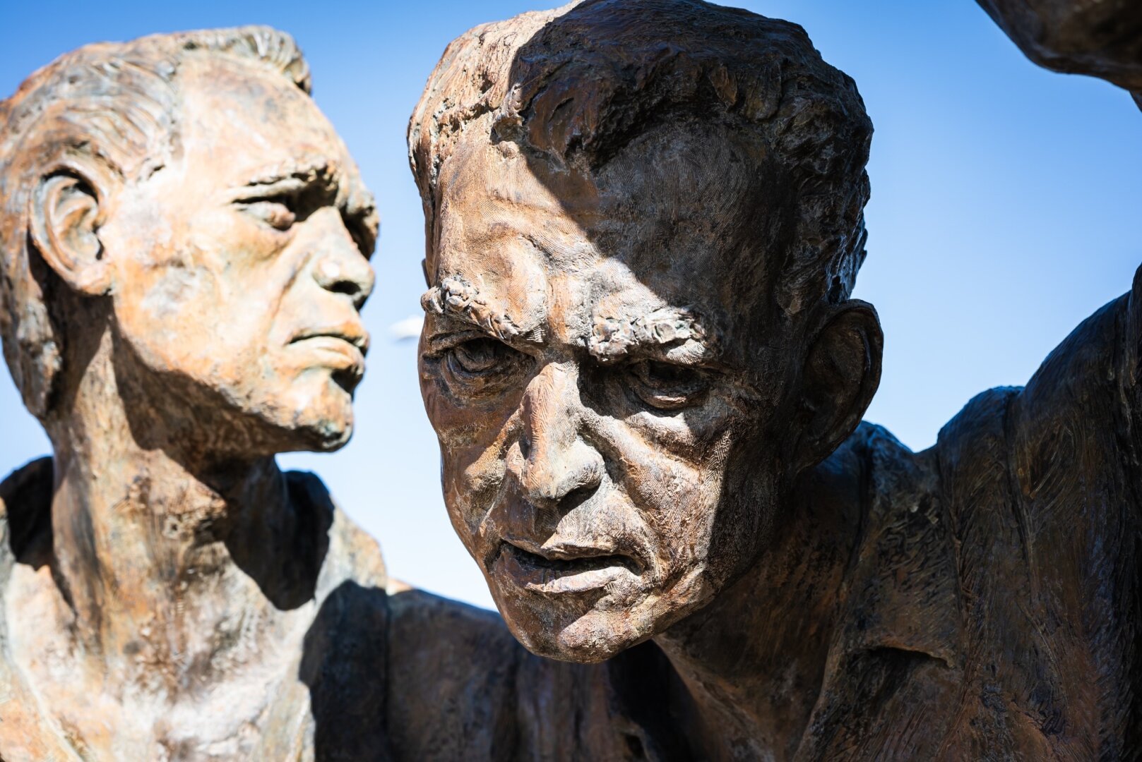 Bronze sculpture of two U.S. military figures, one in a helmet staring ahead with intensity, the other slightly behind with a composed expression, both part of a war memorial set against a clear blue sky.