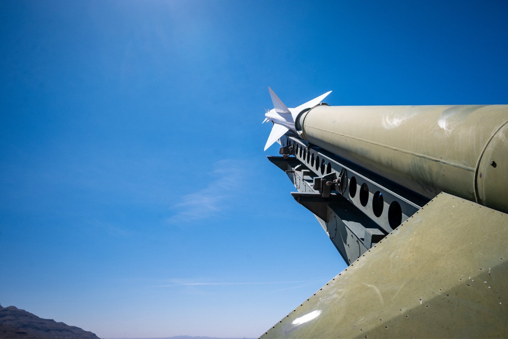 Close-up of a Cold War-era missile launcher pointed at a cloudless blue sky, photographed from below. Desert mountains visible in the far background
