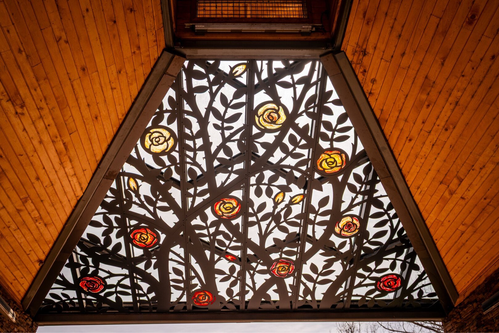 Overhead view of a decorative wooden canopy featuring metal panels with rose cutouts and stained glass inlays, set against a cloudy sky
