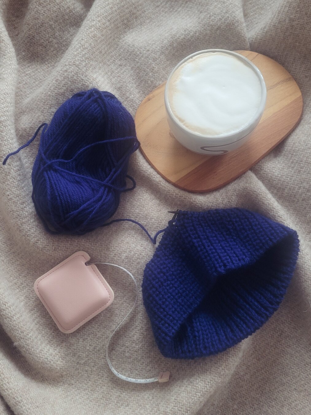 DEU Auf einer beigen Wolldecke sind der Anfang einer Strickmütze aus blauer Wolle sowie ein Maßband und eine Tasse Cappucino zu sehen. 

ENG Displayed on a brown woolen blanket are a not finished handknitted hat in bright blue, a pink tape measure and a cappucino.