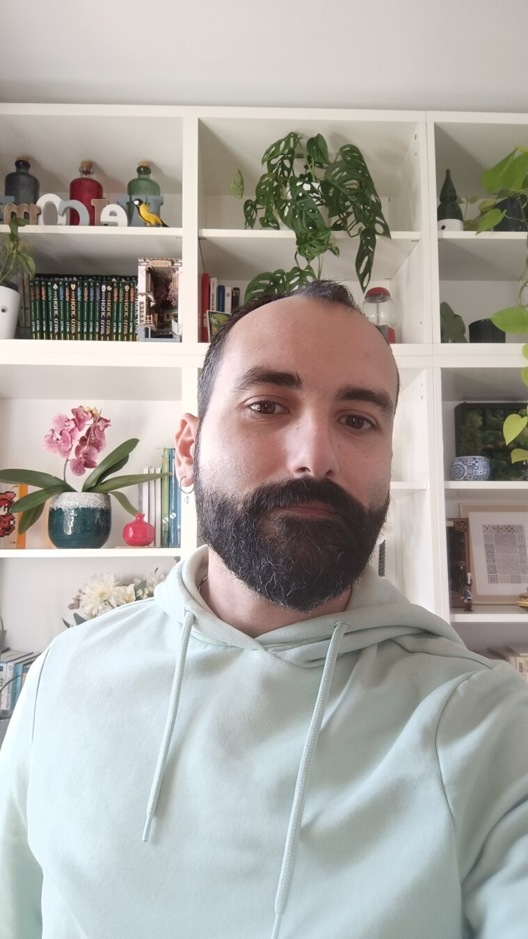 Foto con mi barba. Llevo una sudadera de color turquesa y al fondo se ve mi librería con plantas.