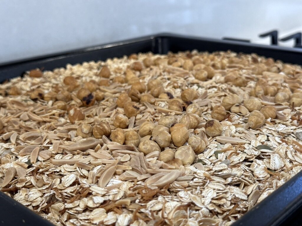 Oven tray showing freshly baked muesli with hazelnuts and sliced almonds toasted on top.