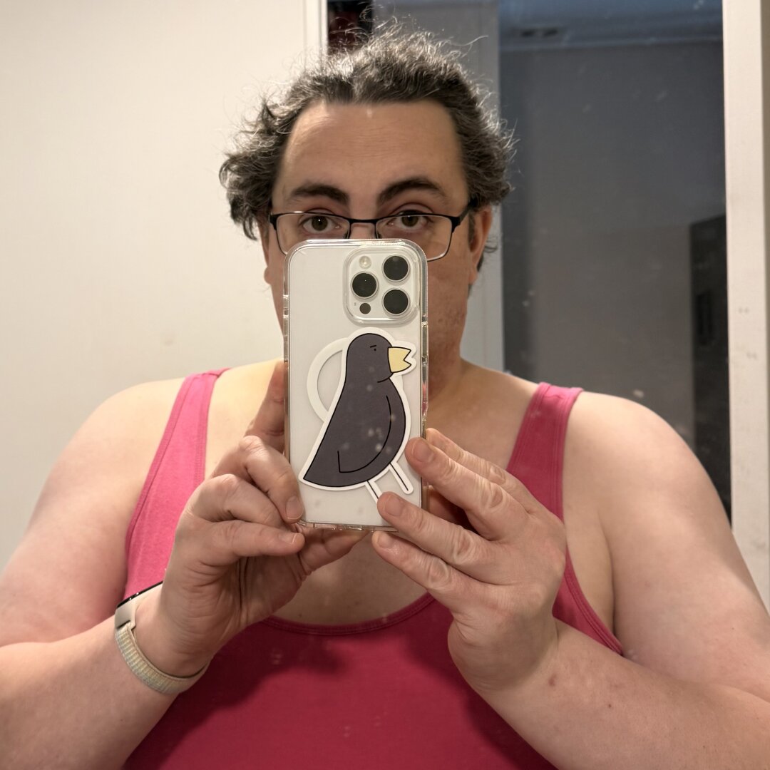 a mirror selfie of me, rebecca swift, in a pink tank top hiding most of my face with my white iPhone 16 Pro Max i a transparent case with a sticker of Bird from Poorly Drawn Lines stuck over most of the magsafe ring.