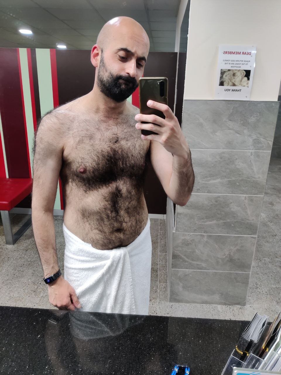 A man taking a selfie in front of a mirror. He's only wearing a towel on his waist, he has a toned body and a very hairy chest, a beard and a shaved head