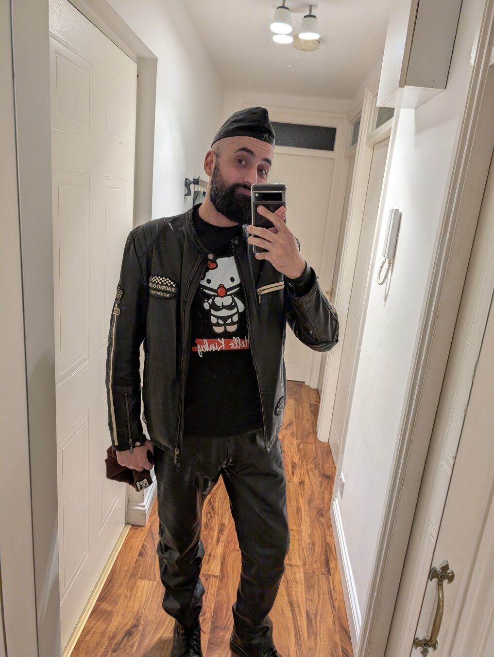 A selfie of a man wearing boots, black leather pants and a jacket, which has 2 white stripes on the arms and a patch, and a garrison cap. The t-shirt has a drawing of Hello Kitty in bondage gear and a gag, and it says Hello Kinky under it