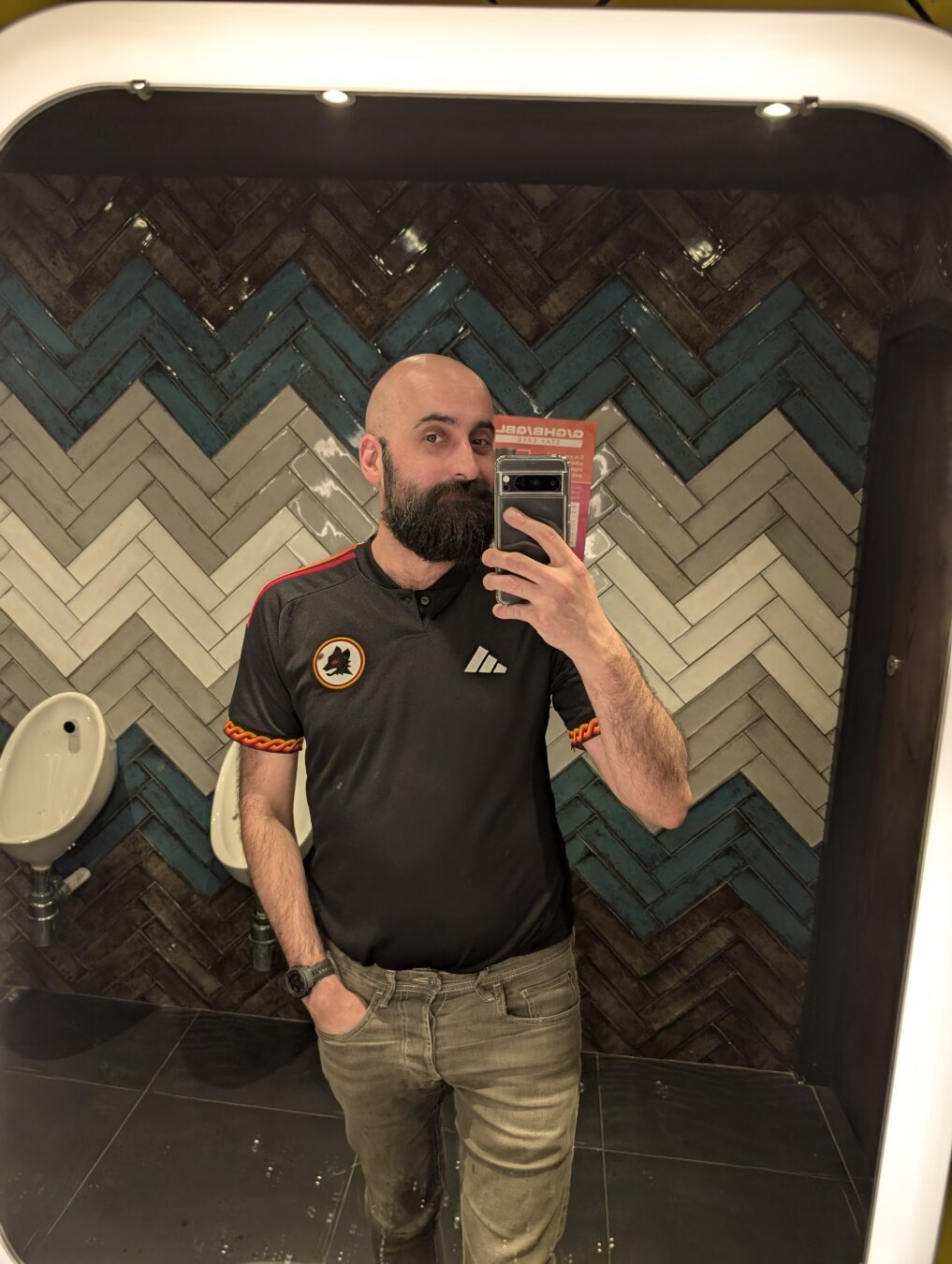 A man taking a selfie in front of a mirror, shaved head and black beard, with a quizzed look on his face. He's wearing a black Roma football t-shirt, and tight jeans.