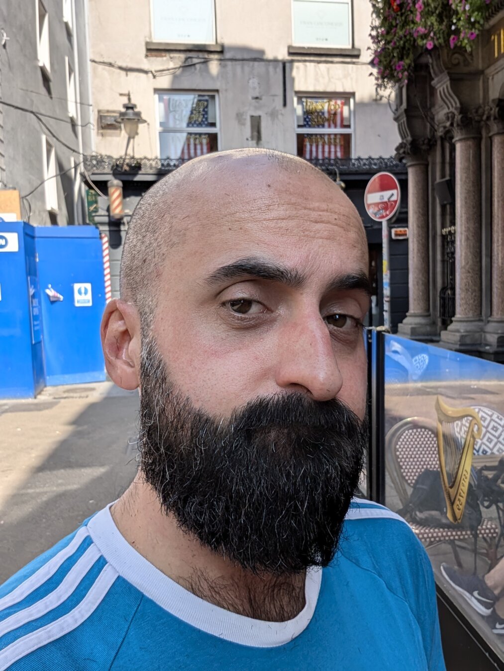 A selfie picture of a man, with a head shaved a couple days before the picture was taken, a thick beard. He's almost doing a concerned bear face. His eyes are brown, and he's wearing a blue Adidas t-shirt. You can see some ches hair coming out of the neck of it