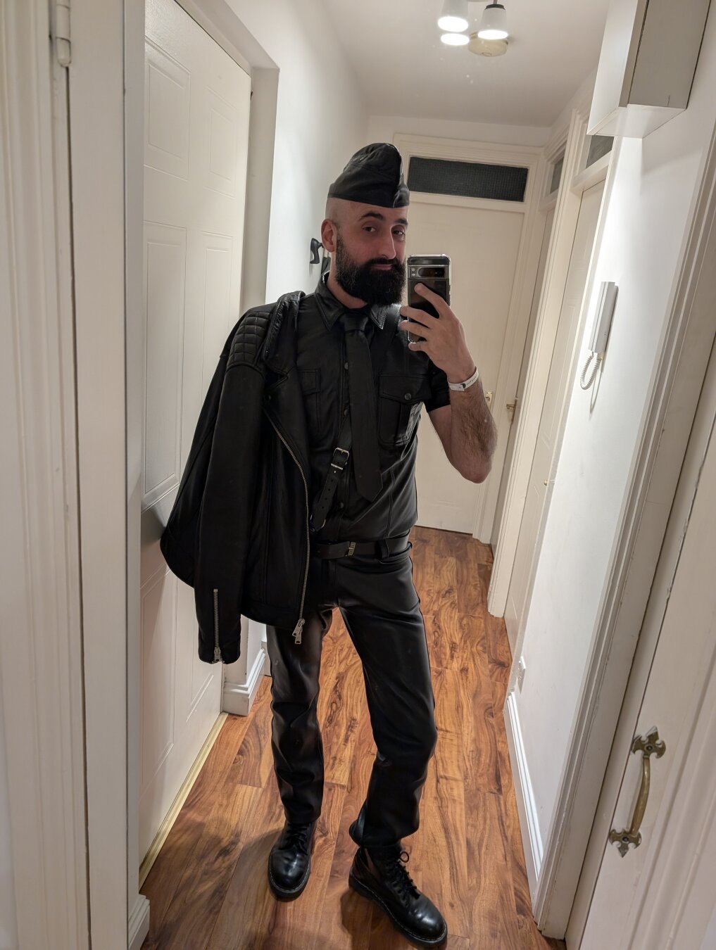 A bearded man taking a selfie, wearing a full black leather outfit, with a jacket on his shoulder