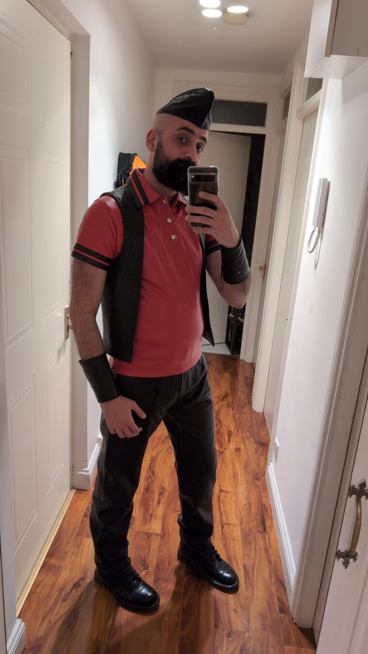 A selfie, a man standing with Dr Martins boots, black leather pants, a red leather polo shirt, a black leather vest, two leather handcuffs and a black leather garrison hat. He has a beard too and a shaved head