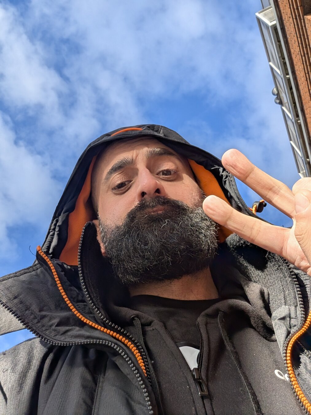 Selfie from the bottom of a bearded man, wearing a black and bright orange hoodie, he's doing a victory symbol with his fingers. In the background you can see a blue sky with some scattered clouds