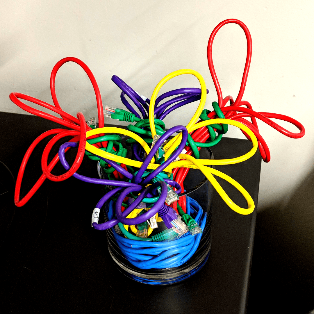 A glass jar on a black table. The jar contains red, yellow, and purple Cat5e network cables arranged in loops to look like a bouquet of flowers. At the bottom there is a blue cable that wraps around the inside of the glass, simulating water.