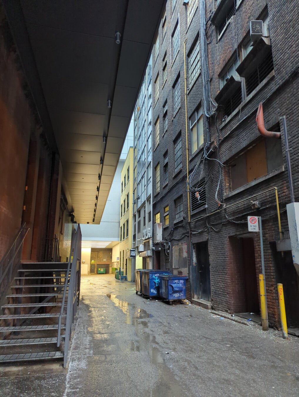 An alley way between two buildings. The ground is wet from rain. On the left is a single brown brick building several meters long with a black overhand. There are corrugated metal stairs rising about one meter to a side entrance. On the right are the soot stained back of several adjoining buildings and loading doors. In front of some of them two blue dumpsters with graffiti. Ad the end of the alley there is an orange lit loading dock area.
