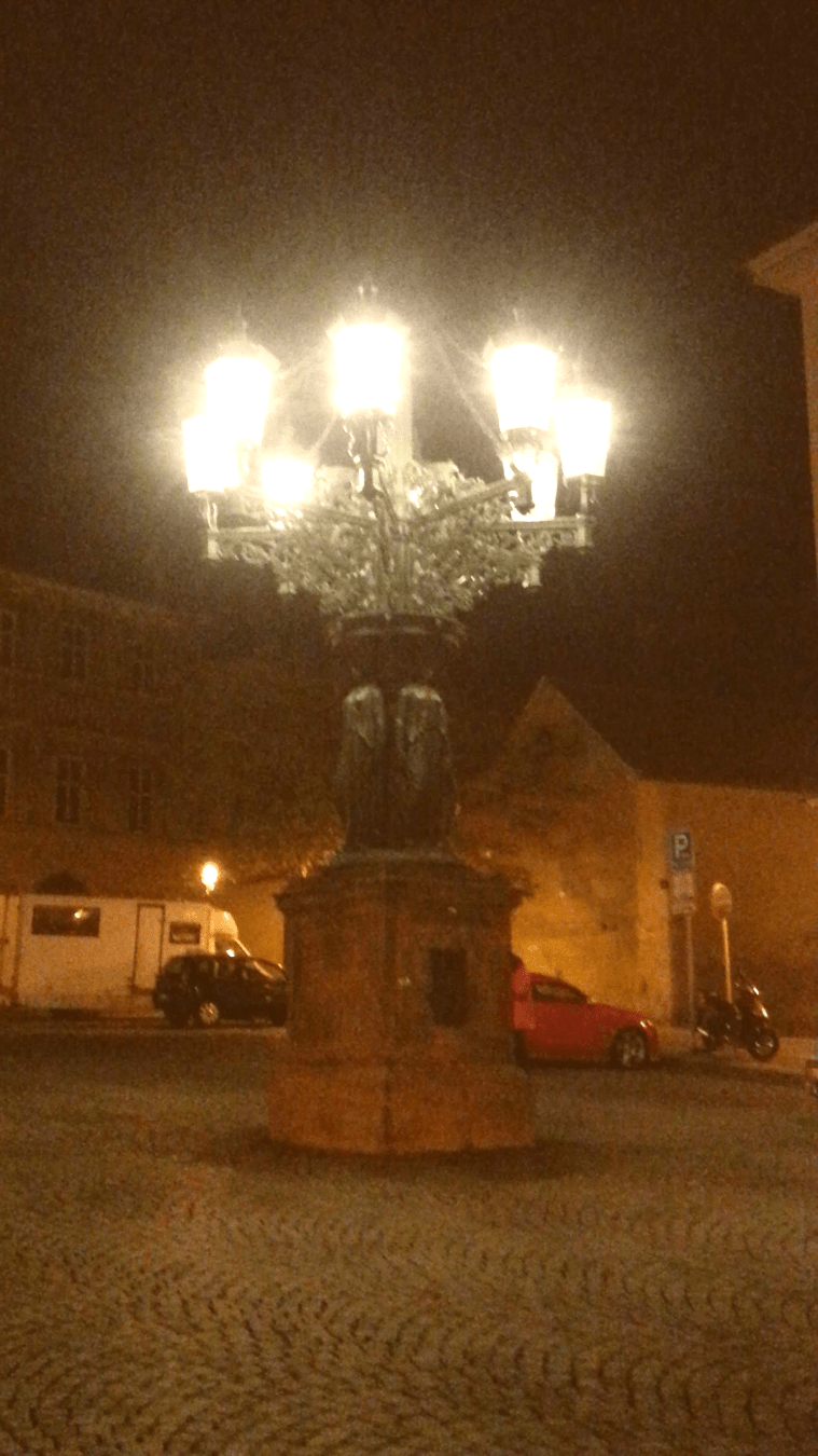 A night time photo of a large 8 lamp Kandelábr in a square in Prague. The lamps are bright white against a dark background. There is a 2 story building in the background lit with orange street lights.