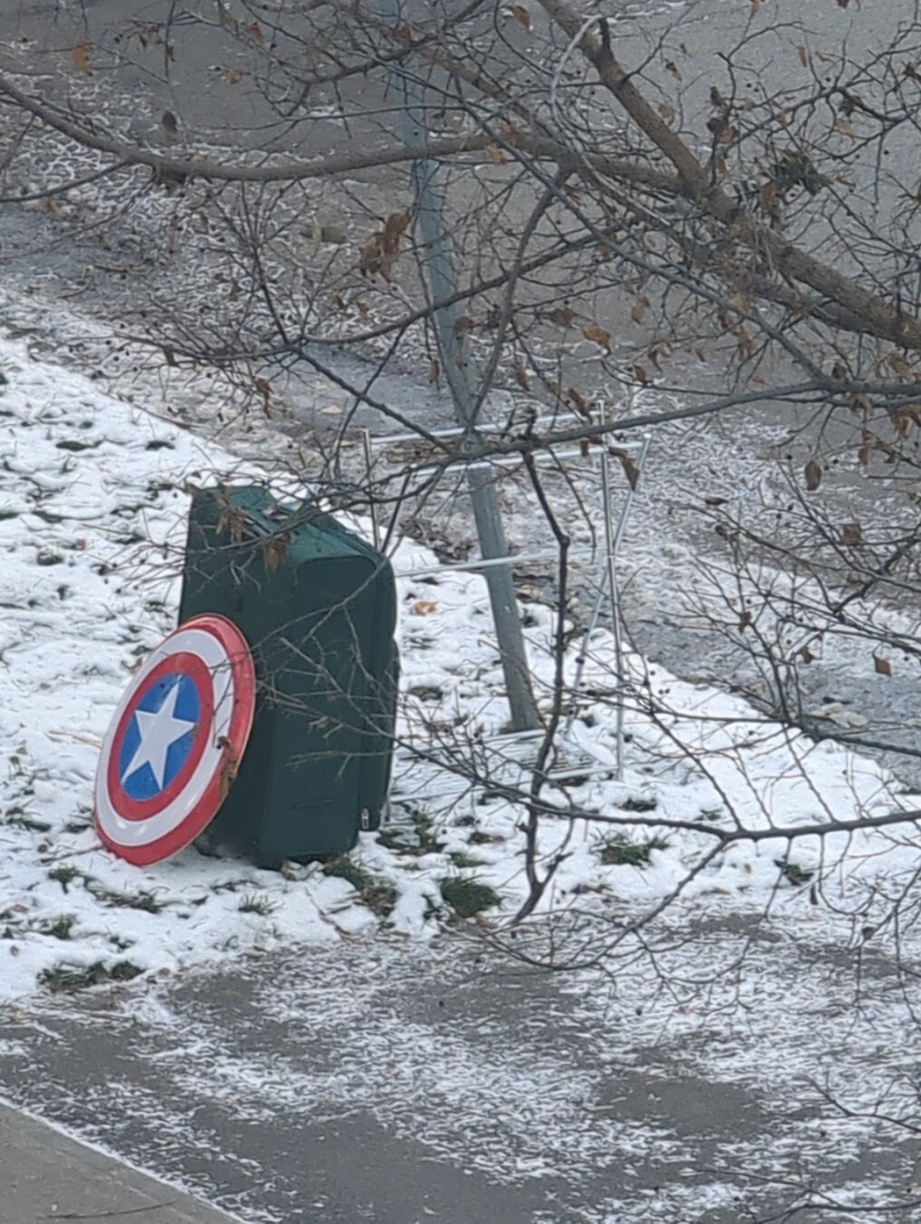 A suitcase sitting on a snowy roadside. Leaning against the shield of captain america - a circular shield with a red ring, white ring, blue centre and white star in the middle.
