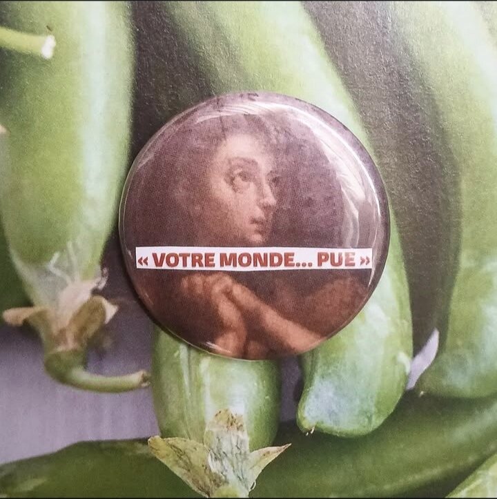 Badge représentant un personnage d'une vieille peinture, probablement religieuse. Le personnage a les yeux et le visage tourné vers le ciel, les mains jointes sous le côté de son visage. Sur lui, le texte 