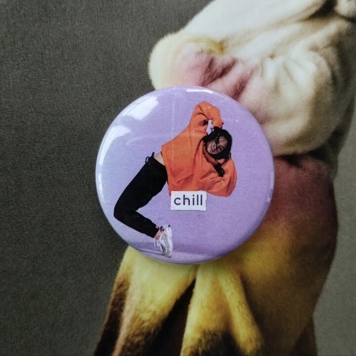 Un badge avec un fond violet. Dessus, une personne dans une position qui a l'air pas pratique et inconfortable : sur la pointe des pieds, penchée vers l'arrière, les mains derrière la tête. Sur l'image, le texte 