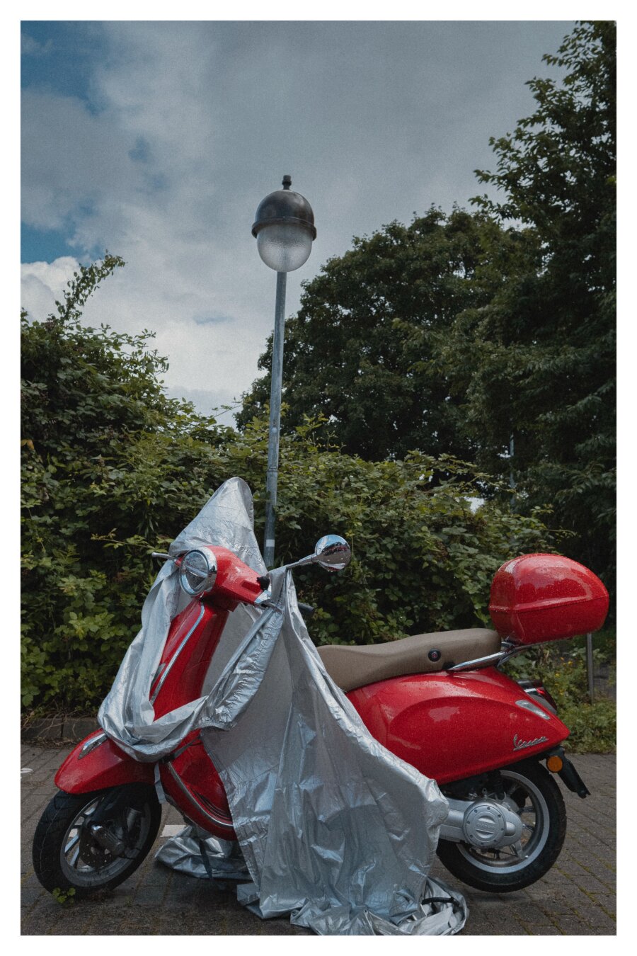 Ein roter Motorroller steht nach dem Regen nass auf einem Parkplatz. Über dem Roller ist ein Regenschutz, der vom Wind halb vom Roller geweht wurde. Im Hintergrund sind Büsche und eine Laterne.