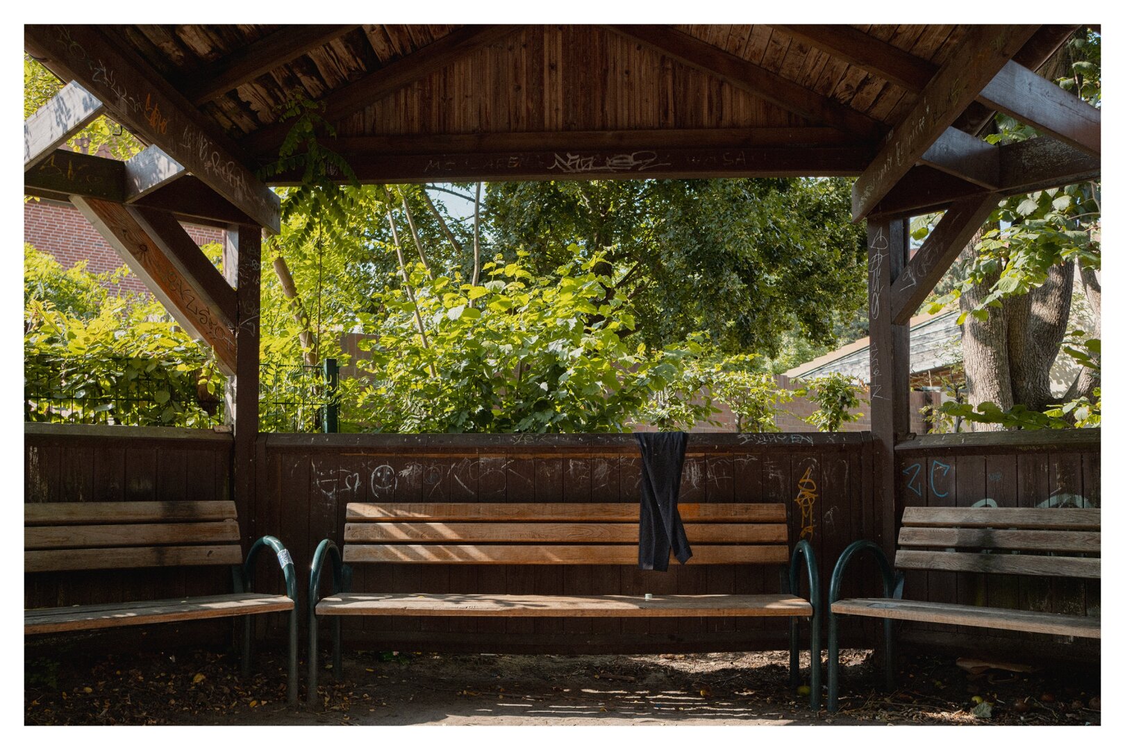 A black trouser is hanging over the edge of a wooden pergola witch benches. In the background there are trees and houses.