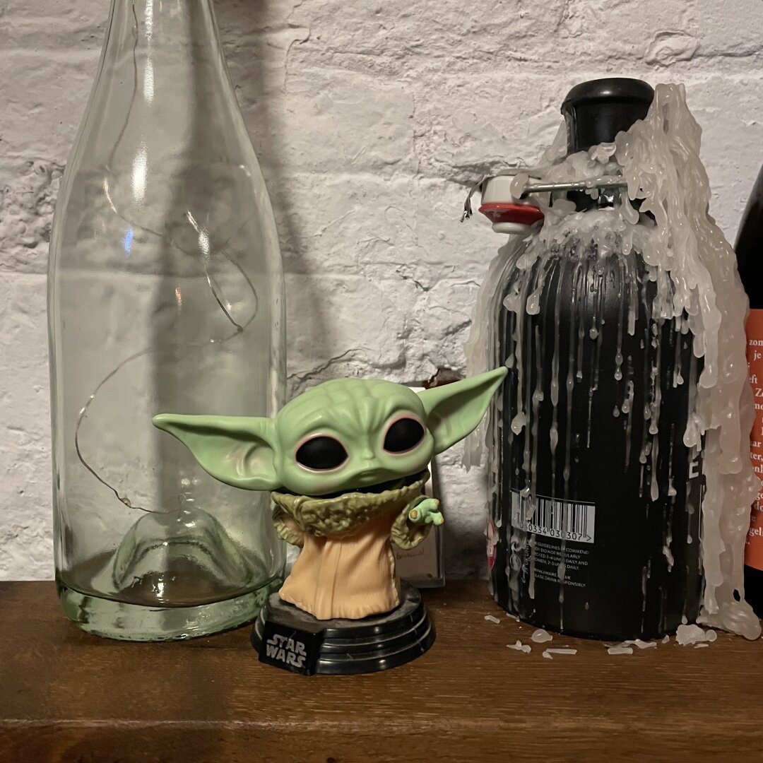 A green coloured figurine of a film character called Grogu stands in front of two glass bottles. One bottle is covered in candle wax.