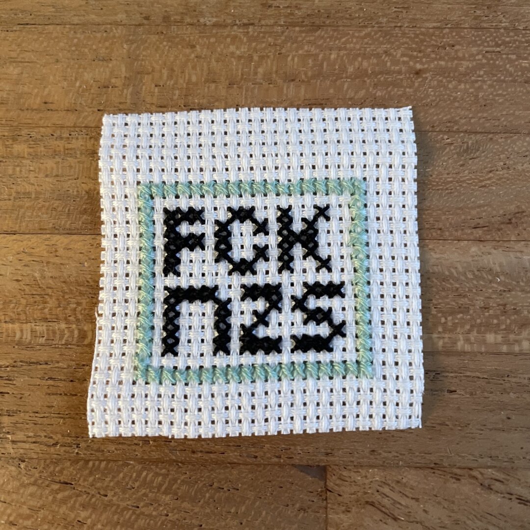 A small cross stitch embroidery piece. There are black letters spelling FCK NZS on white embroidery cloth with a light green border around them.