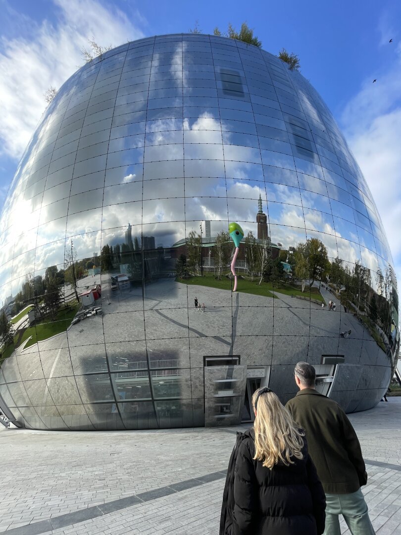 An egg shaped building with the top cut off. The building is made from mirrors. On the outside in the mirrors you can see the skyline of the city of Rotterdam, with blue sky and clouds. In front of the building are a girl with long blonde hair and a man with dark hair, seen from the back. They are looking at the building.