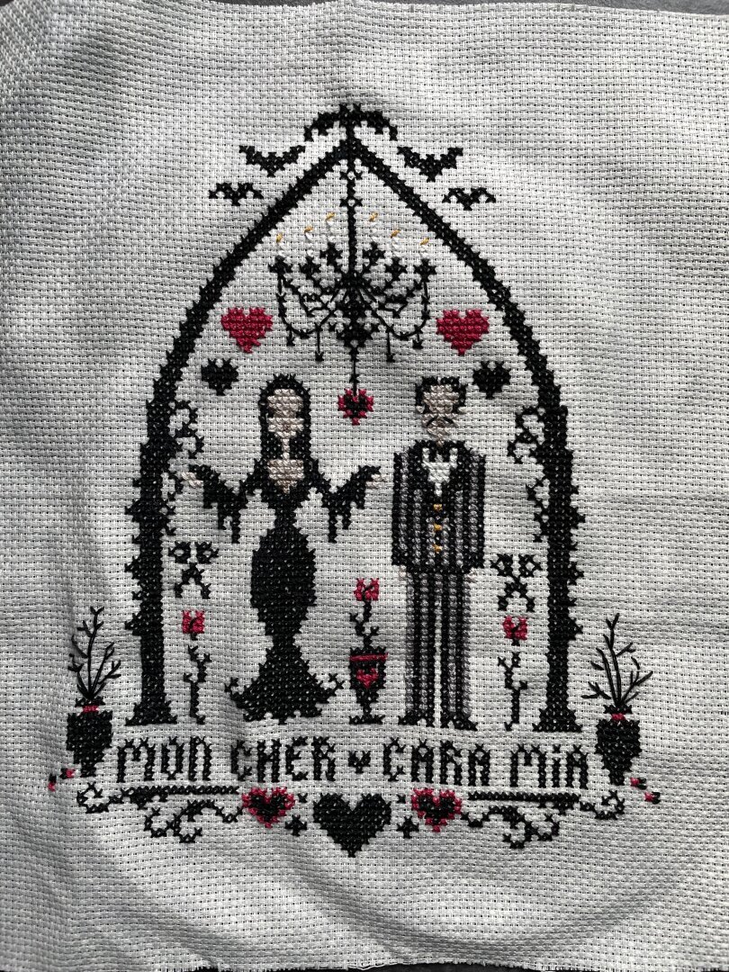 A cross stitch embroidery piece showing Morticia and Gomez Addams under an arch. Besides them are roses and above them is a candelabra. Under them is the text ‘Mon Cher, Cara Mia’.