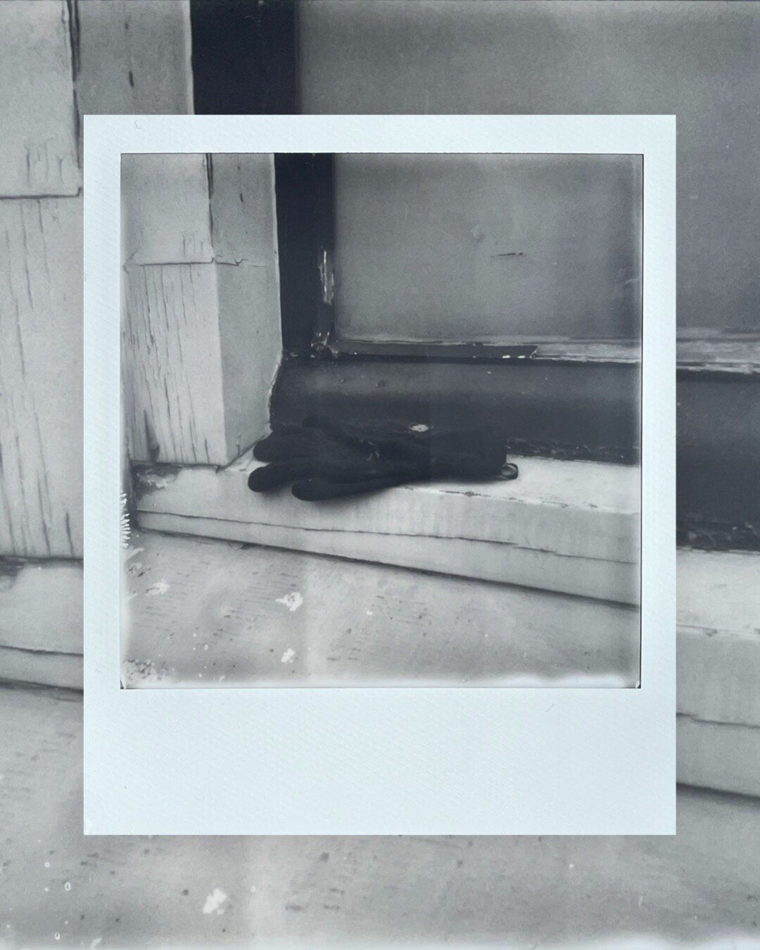 A black and white Polaroid photo of a black glove in a window sill. The paint on the window sill is chipped and the window sill looks weathered.