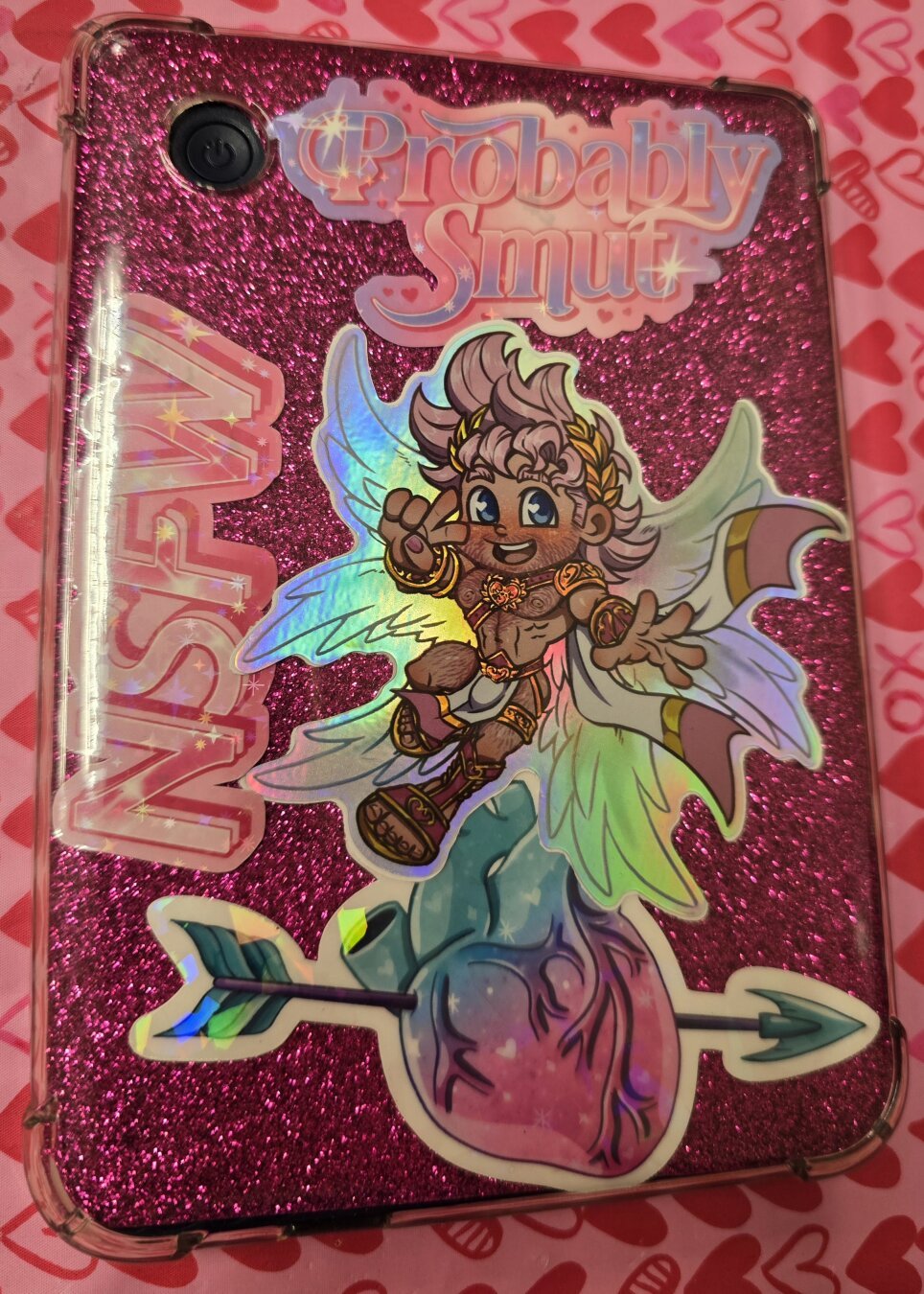 Kobo Clara 2E ereader in a clear case with a pink glitter background with stickers reading "Probably Smut" "NSFW" and a sticker of Eros and one of a heart with an arrow through it