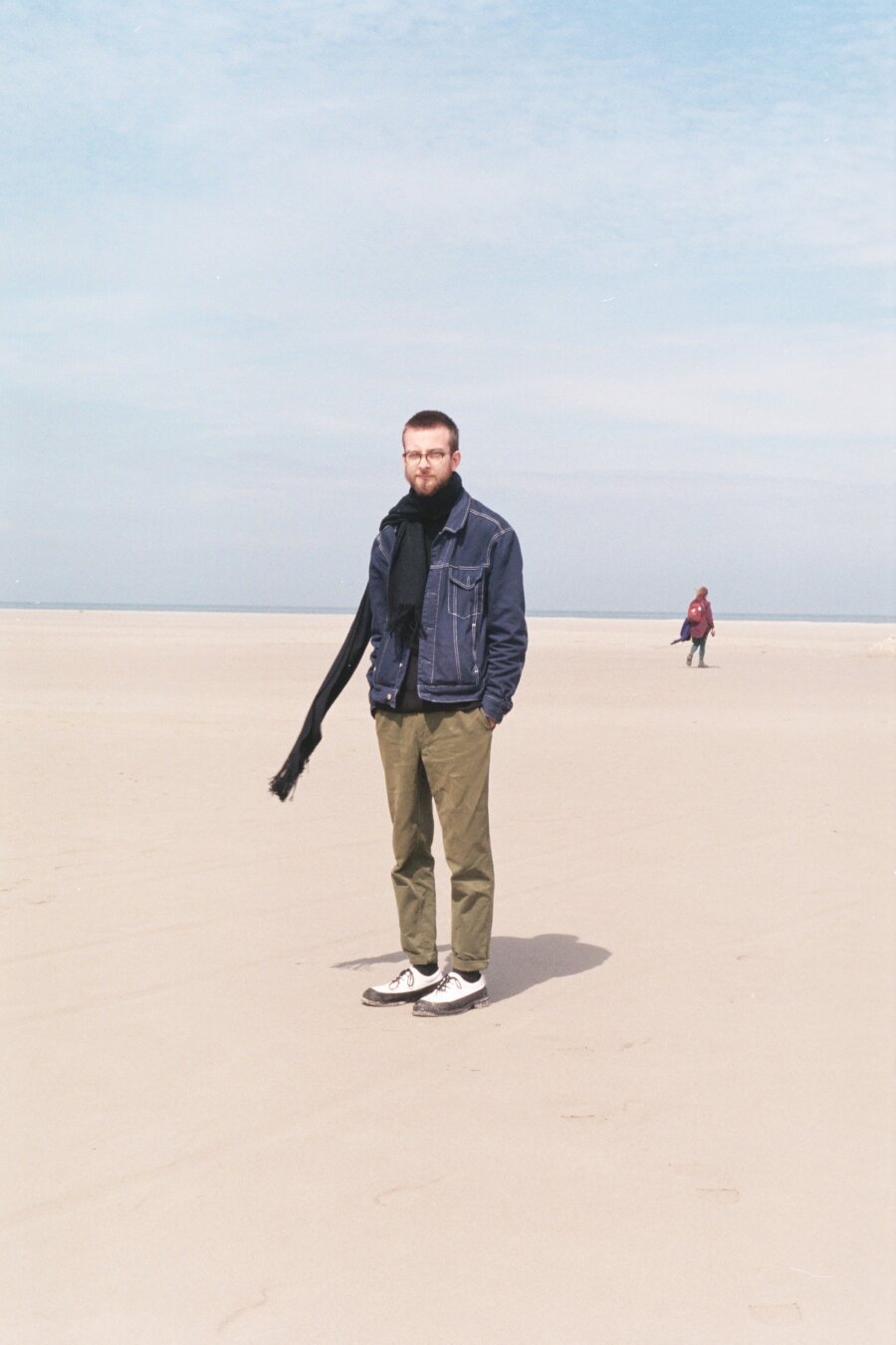 A man wearing a navy blue coat, olive green trousers, black and white shoes and a dark blue scarf is standing in a landscape of seemingly endless sand. The scarf is blowing in the wind. The sky is blue with white fuzzy clouds. In the background on the right is one other person, dressed in shades of red, walking away from the viewer.