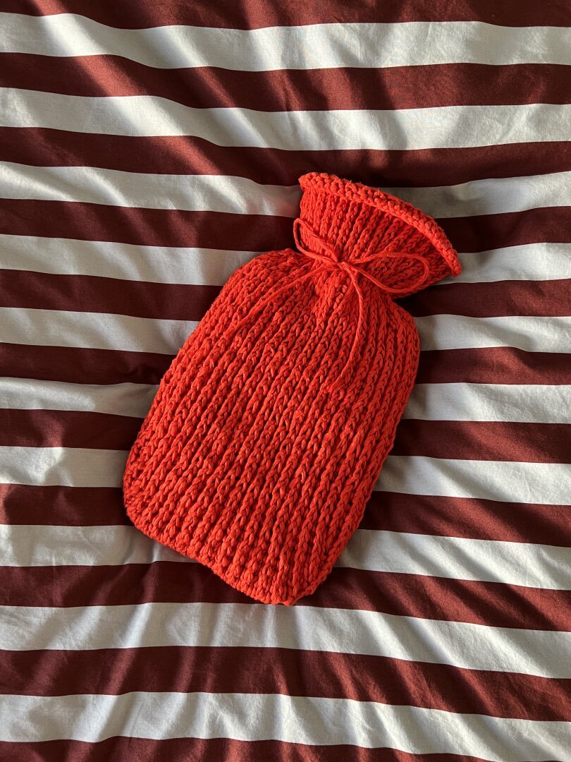 Hot water bottle with a bright red knitted sleeve laying on bedsheets with burgundy and light blue stripes.