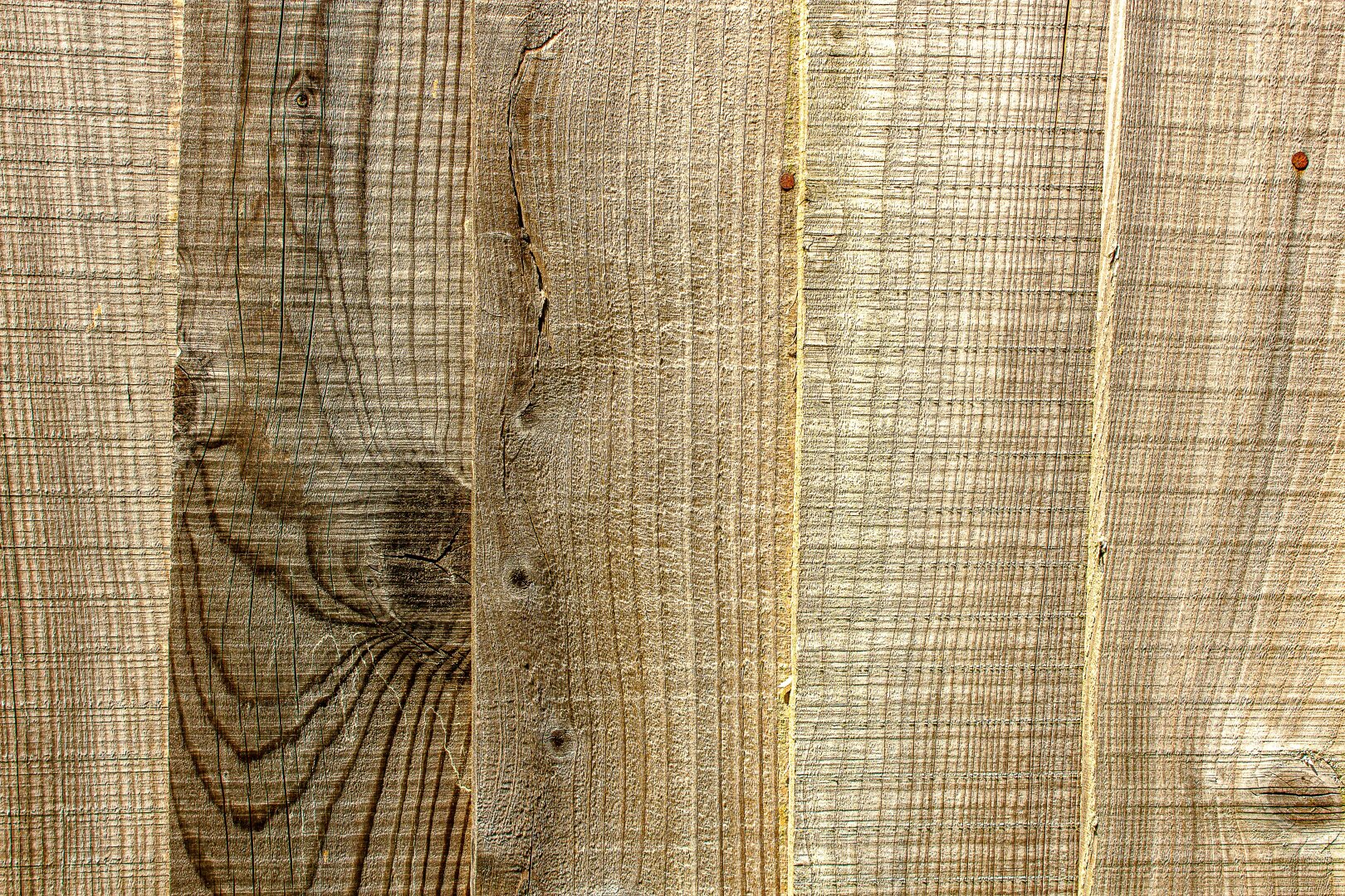 Close‑up of a wooden fence made of vertical planks with visible grain patterns, knots, and fine saw marks. The wood appears rough and weathered, showing natural variations in tone from light tan to medium brown, with two small rusty nails near the top securing the boards.