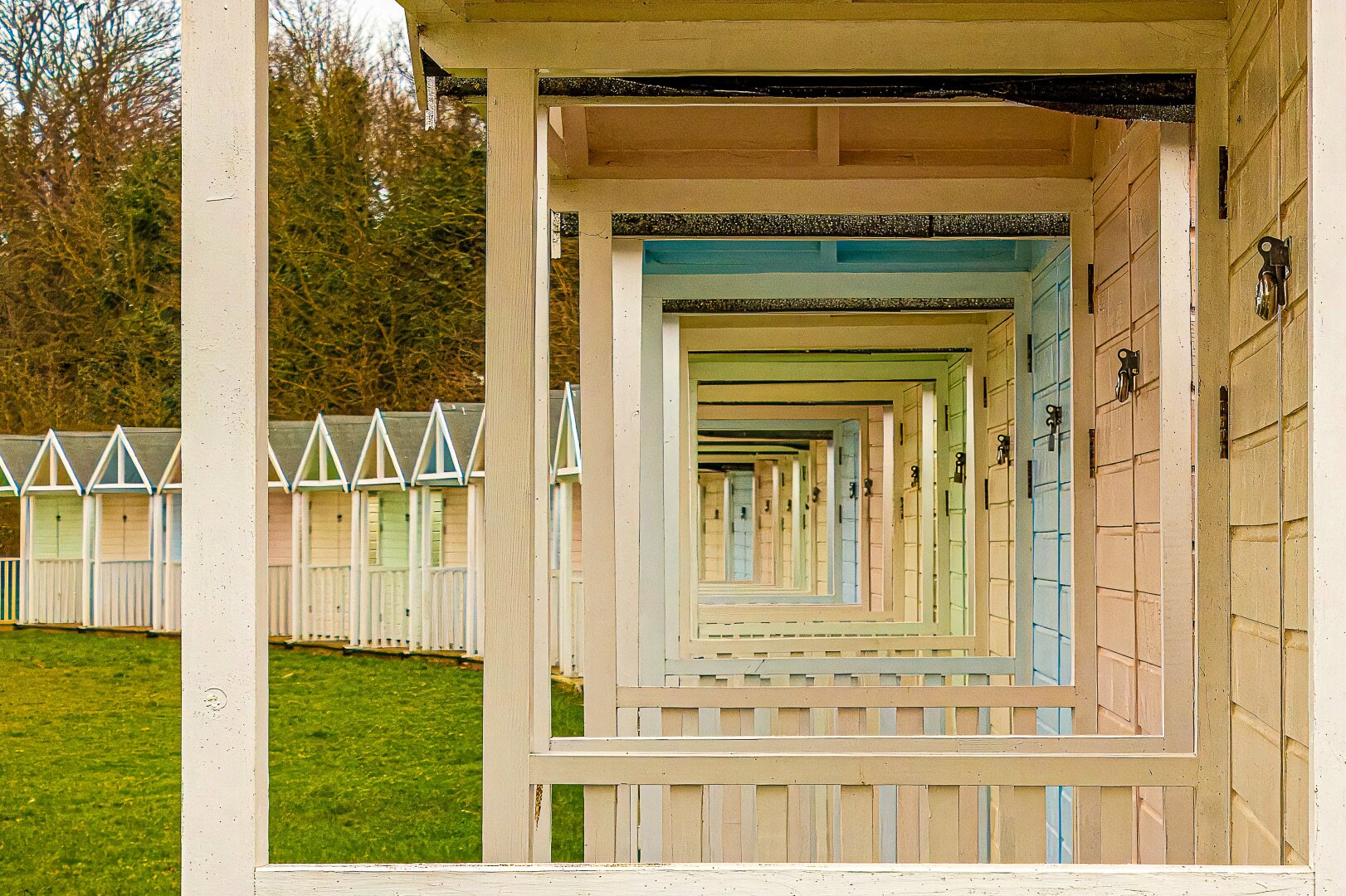 A row of pastel‑coloured beach huts in Devon viewed through their open front frames, creating a tunnel‑like perspective. The repeating rectangular structures form a geometric pattern that draws the eye inward, with soft hues of cream, blue, green, and pink adding rhythm and depth against a grassy backdrop.