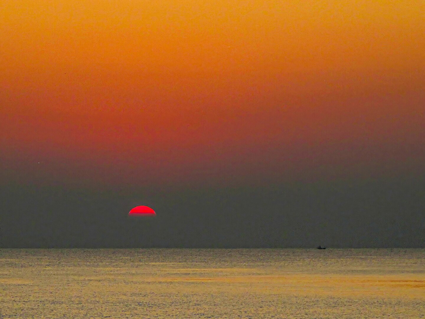 Sunrise over the sea at Aleenta, Pranburi, Thailand. The deep red sun sits half above the horizon, casting a warm gradient of orange and gold across the sky and reflecting softly on the calm water. A small boat silhouette appears in the distance, adding scale to the tranquil scene.