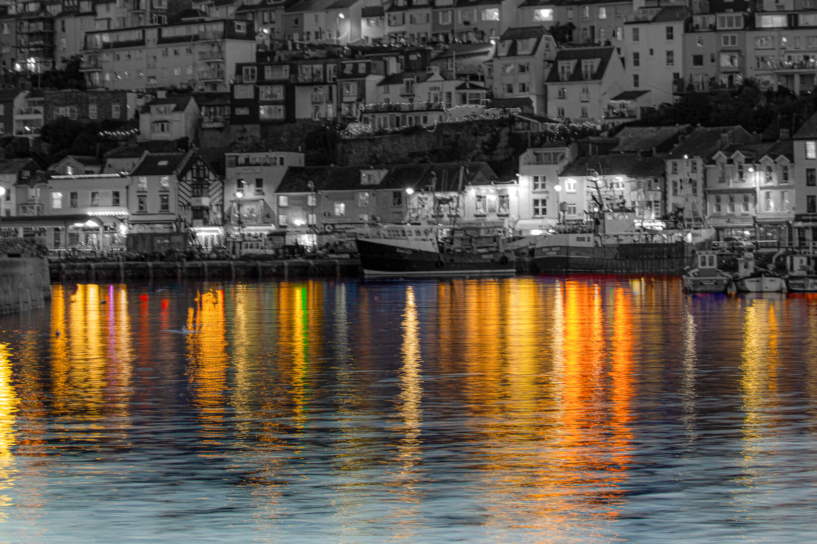 Night view of Brixham Harbour with colourful reflections of yellow, orange, red, and green lights shimmering on calm water. The hillside town behind is shown in black and white, with boats moored along the quay and houses stacked above, creating a vivid contrast between monochrome buildings and glowing harbour reflections