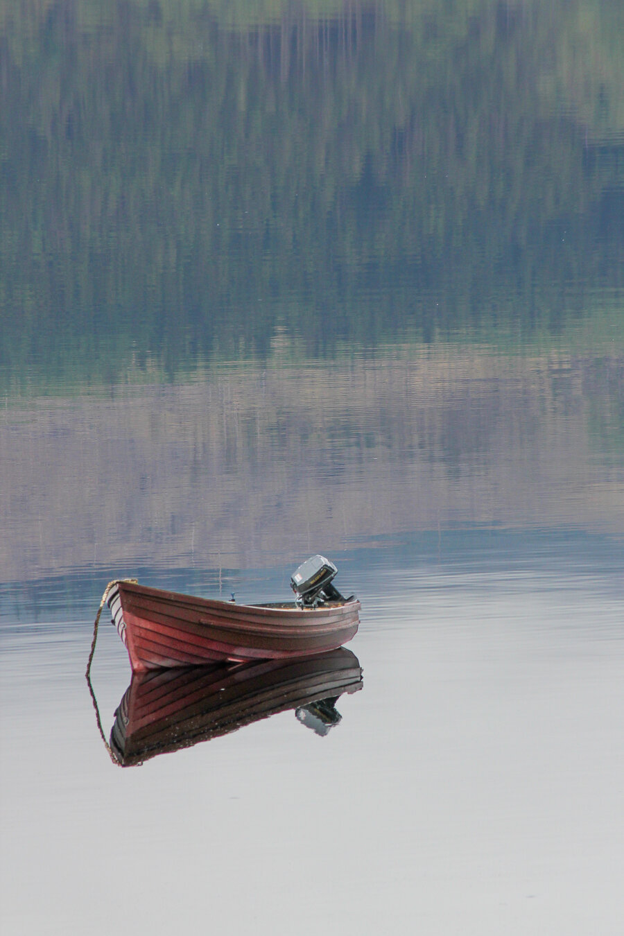 A small wooden boat with an outboard motor floats motionless on the glassy surface of Loch Fyne in Argyll, Scotland. The calm water mirrors the boat and the soft, muted reflection of forested hills, creating a tranquil, minimalist composition.