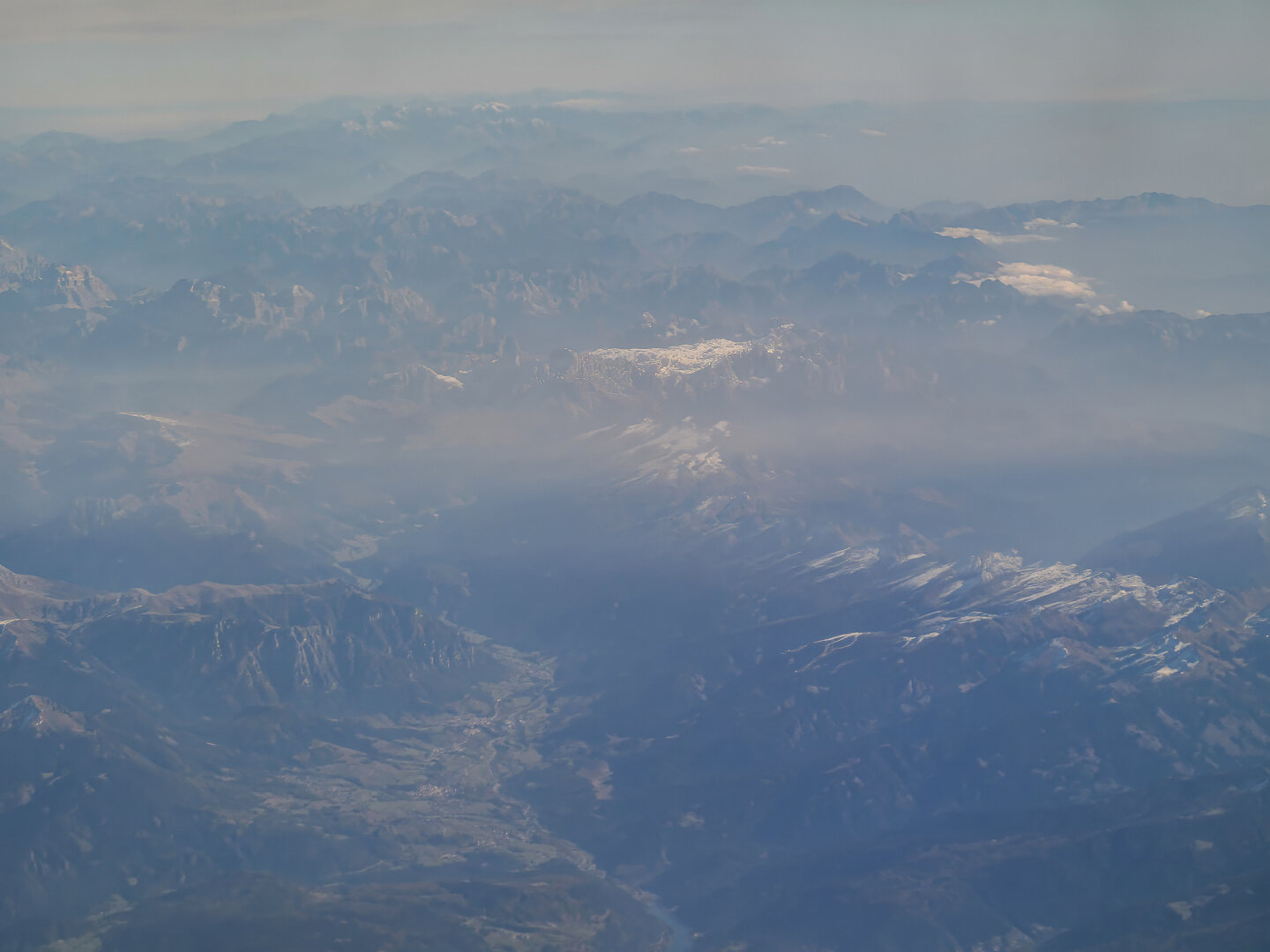 Aerial view of the Alps bathed in soft early‑morning light, with hazy blue ridges fading into the distance and snow‑tipped peaks glinting under a pale sky.
