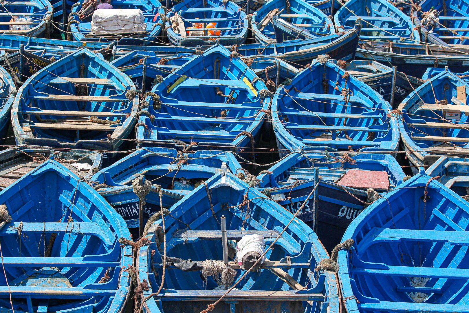 A dense cluster of small wooden fishing boats painted in vivid blue fills the frame at the harbor in Essaouira, Morocco. The boats are tightly packed and secured with weathered ropes, creating a striking geometric pattern of overlapping hulls and lines. Sunlight highlights the worn paint and textures of the wood, evoking the daily rhythm of coastal life.