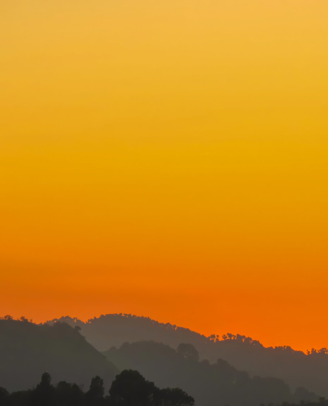 Sunset over the Margalla Hills in Islamabad, Pakistan. Silhouetted ridges and trees form dark layers beneath a vivid sky that shifts from deep orange near the horizon to golden yellow above, creating a tranquil gradient of evening light.