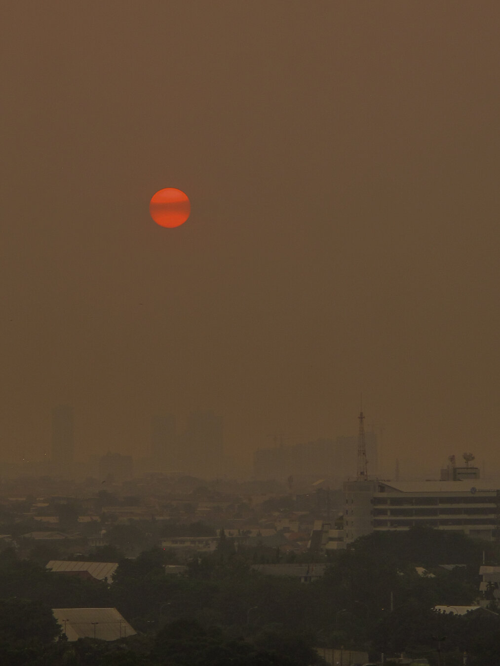 Reddish‑orange sun rising through thick brown smog over Jakarta, Indonesia, with hazy outlines of high‑rise buildings and rooftops barely visible beneath the polluted sky.