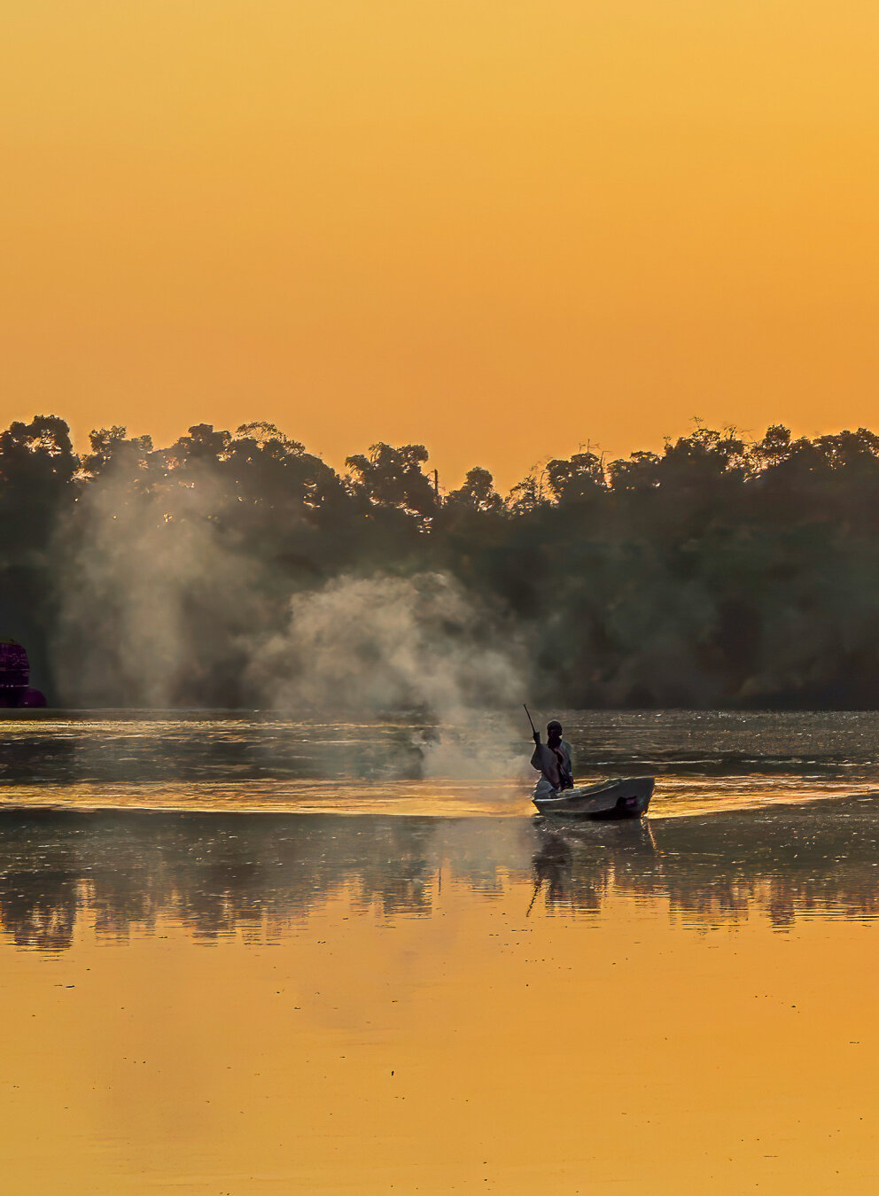A fisherman glides home across the Makasutu River at dawn, his small boat cutting through mist that rises from the golden water. The sky glows amber above the dark silhouette of forest trees, and the still surface mirrors the warmth of morning light. The scene feels timeless — a quiet rhythm between human and nature at the edge of day.
