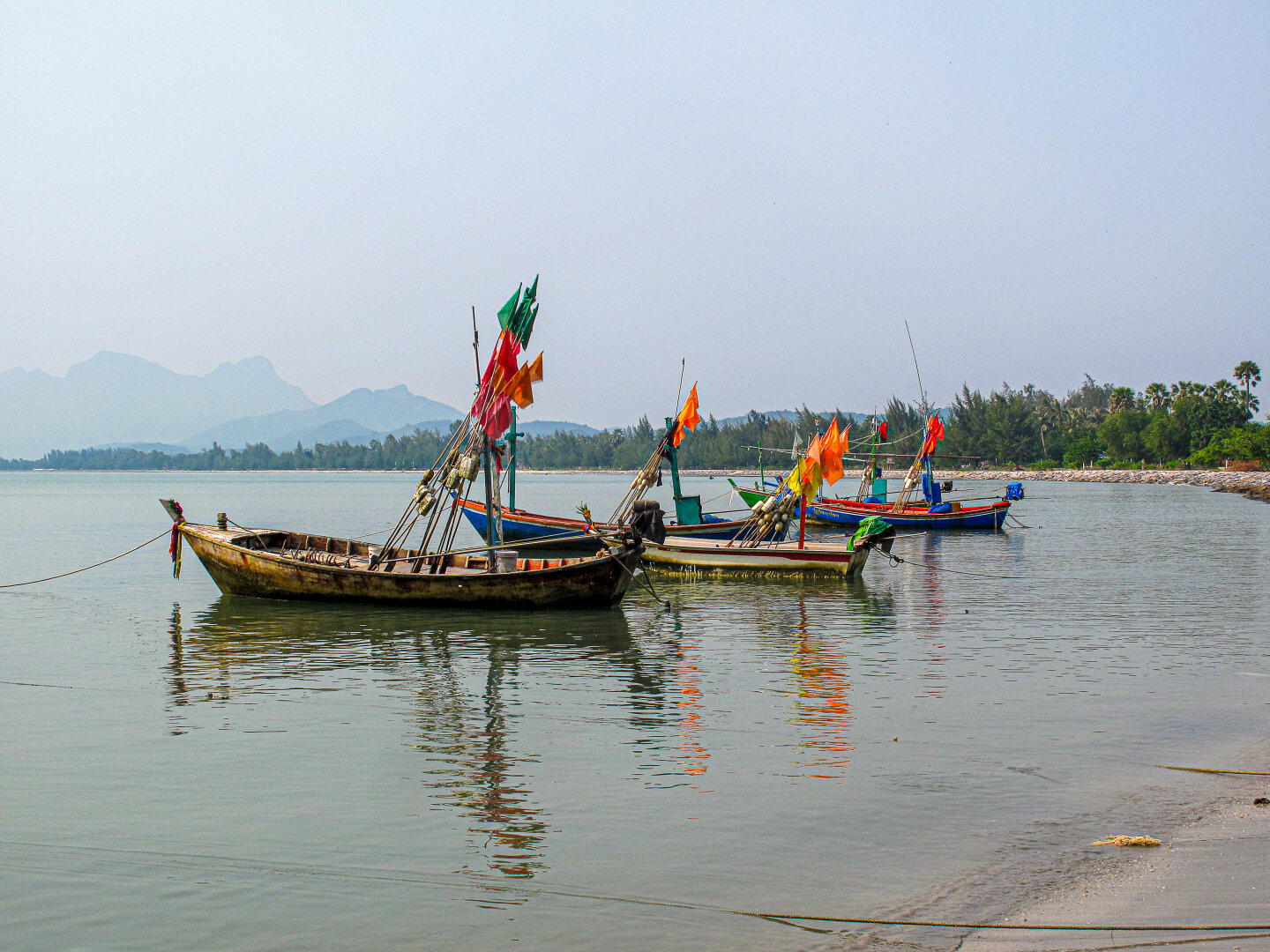 hree moored wooden fishing boats with colorful flags on calm water at Pranburi, Thailand, with hazy hills beyond