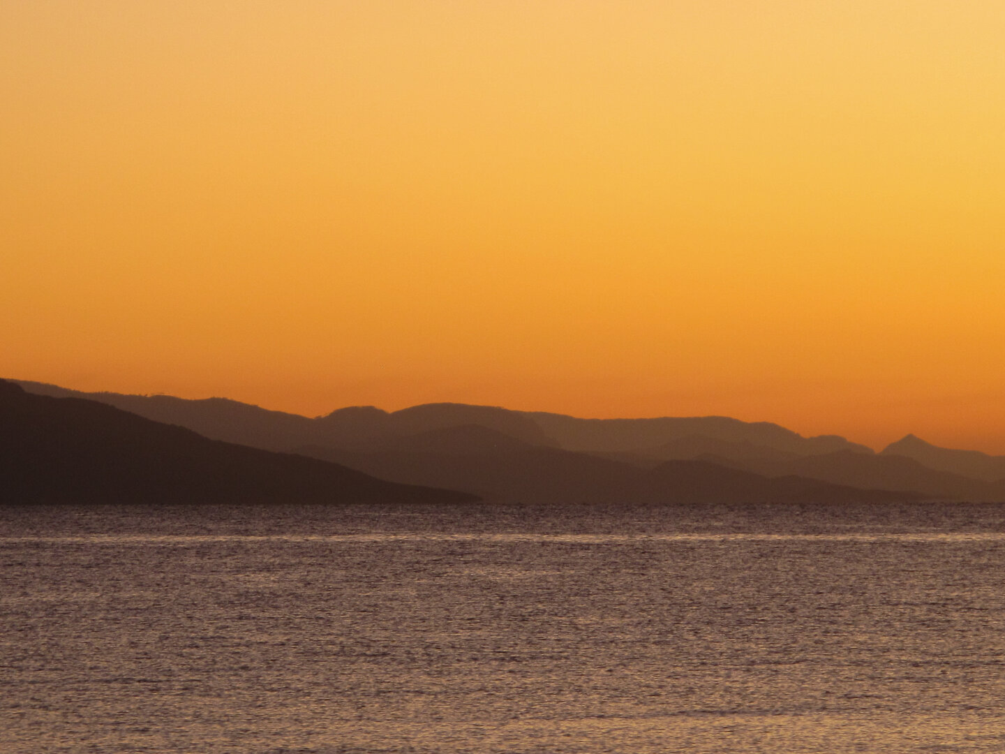 Golden dawn light glows over the Turkish coast seen from Kos, Greece, with calm sea water in the foreground and layered mountain silhouettes fading into the warm orange sky.