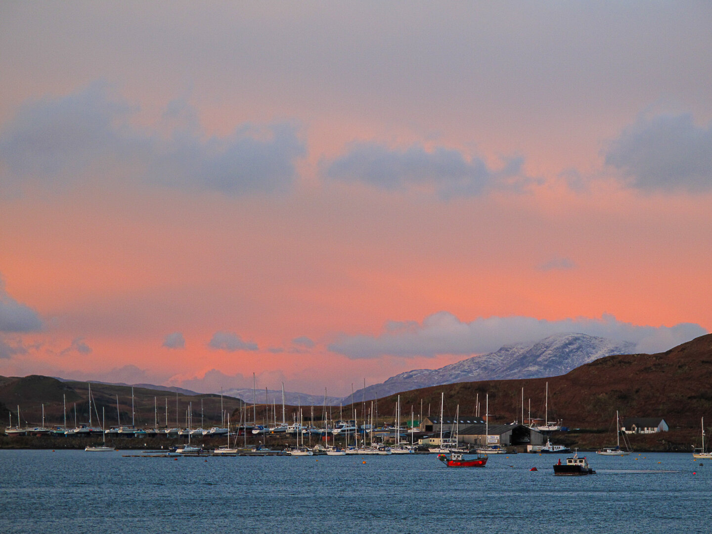 Ben More on the Isle of Mull seen from Oban, Argyll, Scotland at sunset. The harbor is dotted with moored sailboats and small vessels, their masts silhouetted against a soft pink and orange sky. Snow‑tipped mountains rise in the distance beneath scattered clouds, and the calm water reflects the evening light.